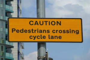 Gelbes Warnschild an einem Pfahl vor einem Gebäude, mit einem klaren blauen Himmel im Hintergrund, auf dem steht "Fußgänger überqueren Fahrradspur".
