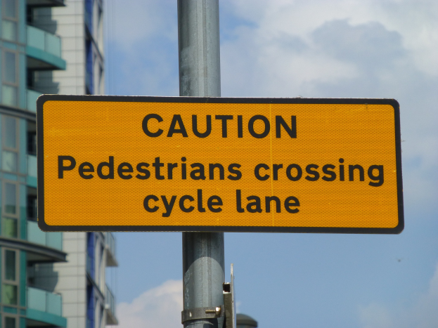 Gelbes Warnschild an einem Pfahl vor einem Gebäude, mit einem klaren blauen Himmel im Hintergrund, auf dem steht "Fußgänger überqueren Fahrradspur".