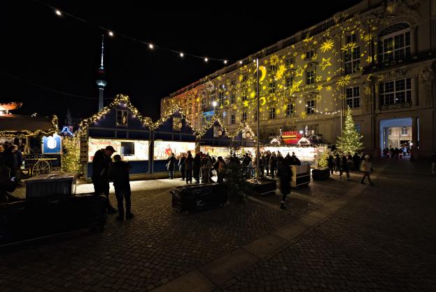 Ein belebter Weihnachtsmarkt in Berlin, Deutschland mit Menschen um dekorierte Stände, festliche Lichter und Gebäude im Hintergrund unter einem dunklen Himmel.