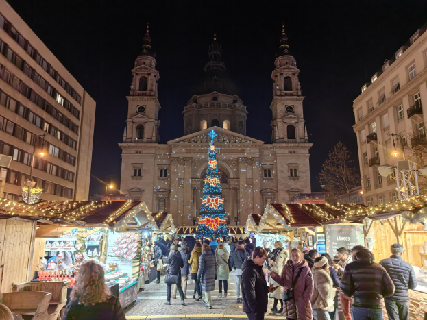 Weihnachtsmarkt mit festlichen Ständen, Lichtern und Menschen vor einer Kirche bei Nacht, mit Gebäuden, Bäumen und einem sternenklaren Himmel im Hintergrund.