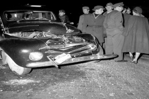 Eine schwarz-weisse Szene eines beschädigten Autos am Strassenrand, mit einer Gruppe von Menschen drumherum, die das Auto besorgt anschauen.