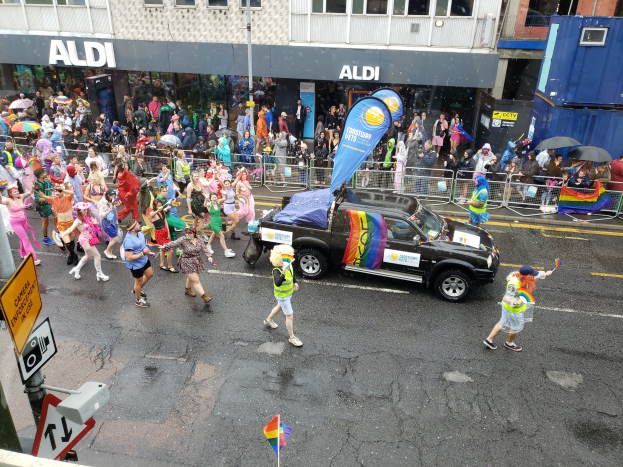 Eine Gruppe von Menschen in bunter Kleidung, die eine Straßenparade entlanggehen, mit einem Auto in der Mitte, einige halten Schirme, und Gebäude mit Fenstern im Hintergrund.