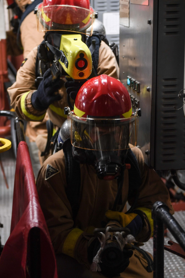 Feuerwehrleute in Schutzausrüstung, einer hält einen Feuerlöscher, mit einem Metallobjekt und einem roten Tuch im Hintergrund.