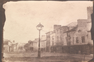 Altes Schwarz-Weiß-Foto einer Straßenlaterne an einer Kreuzung, flankiert von mehrstöckigen Gebäuden unter einem wolkenlosen Himmel.