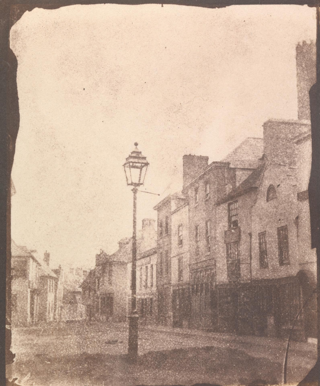 Altes Schwarz-Weiß-Foto einer Straßenlaterne an einer Kreuzung, flankiert von mehrstöckigen Gebäuden unter einem wolkenlosen Himmel.