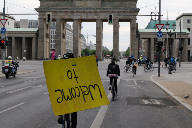 Eine Gruppe von Menschen in Helmen fährt auf Fahrrädern eine Straße entlang vor dem Brandenburger Tor in Berlin, Deutschland, mit einer Person, die eine gelbe Tafel hält, Laternenpfählen, Verkehrszeichen, Gebäuden, Bäumen und einem klaren blauen Himmel im Hintergrund.