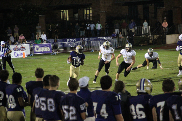 Gruppe von Menschen beim Fußballspielen auf einem Rasenfeld, einige tragen Helme, mit einem Gebäude, Bannern, Treppen, Geländern und Bäumen im Hintergrund.