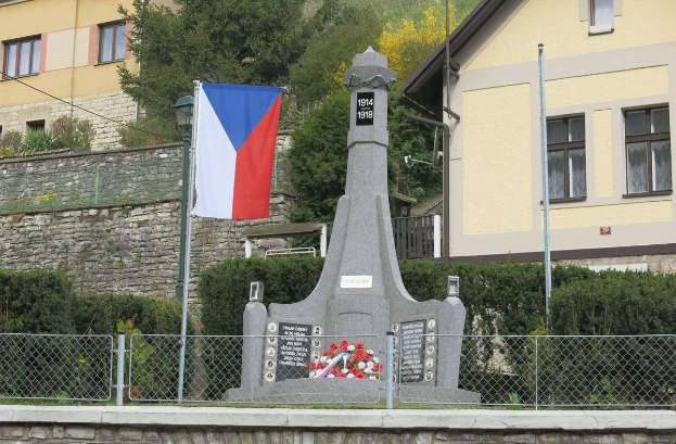 Ein Denkmal mit einer Fahne darauf, umgeben von einem Zaun, Pflanzen, Bäumen und einer Wand, vor einem Gebäude, gewidmet denjenigen, die ihr Leben während des Zweiten Weltkriegs verloren haben.