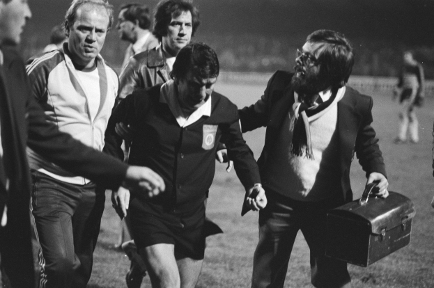 Schwarzes und weißes Bild einer Gruppe von Männern, die auf einem Fußballfeld gehen, einer hält eine Aktentasche, mit offensichtlichem Unbehagen.