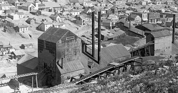 Schwarz-weiß-Foto einer Stadt mit zahlreichen Gebäuden, Häusern, Strommasten, Bäumen, Pflanzen und einer Eisenbahnschiene, die wahrscheinlich ein Kohlebergwerk mit der Stadtlandschaft im Hintergrund zeigt.
