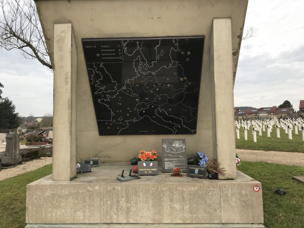 Grabmal eines deutschen Soldaten in einem Friedhof mit einer Karte, umgeben von Blumen und Gegenständen, mit Gräbern, Bäumen, Gebäuden und einem klaren Himmel im Hintergrund.