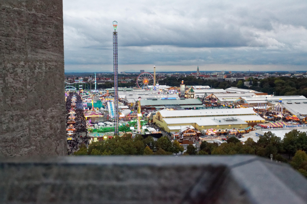 Sicht auf einen Vergnügungspark von oben, mit einer Wand links, Bäumen, Gebäuden, Fahrgeschäften, Pfählen und einem bewölkten Himmel im Hintergrund.