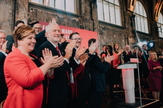 Eine Gruppe von Menschen, die vor einem Publikum applaudieren, mit einem Podium, einem Mikrofon und einer Texttafel auf der rechten Seite sowie Stühlen, einer Fahne, einer Wand, Fenstern und Lichtern im Hintergrund.