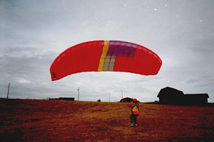 Eine Person mit einem roten Fallschirm segelt in einem Feld, umgeben von Häusern, Pfählen und einem klaren blauen Himmel.