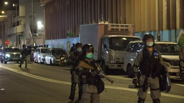 Eine Gruppe von Polizisten in Helmen, Handschuhen und Gesichtsmasken steht nachts auf einer Straße, einige halten Waffen, mit Fahrzeugen, Laternenmasten, Schildern und Gebäuden im Hintergrund.