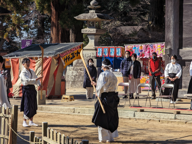Eine Gruppe von Menschen in traditioneller Kleidung steht im Freien in Kyoto, einige tragen Masken und halten Holzstöcke, mit Stühlen, Bannern, einem Zelt und einem hölzernen Zaun unter einem klaren blauen Himmel.