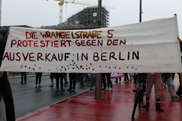 Gruppe von Menschen mit einem Transparent "Die Wrangellstraße 5 Protestiert Gegen den Ausverkauf in Berlin" auf einer Stadtstraße mit Fahrrädern, Pfählen, Gebäuden und einem Kran im Hintergrund.