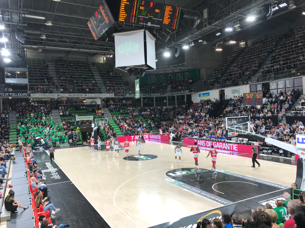 Ein Basketballspiel in einer großen Arena mit Zuschauern auf den Tribünen und Anzeigetafeln, die "Asvel vs Galatasaray" anzeigen, unter einer Decke mit Lampen und einem Bildschirm.