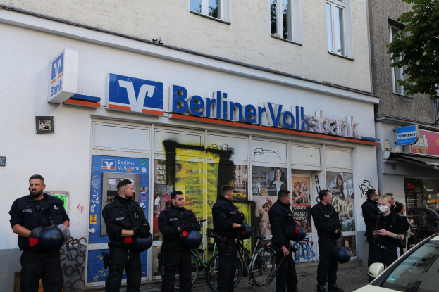 Gruppe von Polizisten vor einer Berliner Volksbank, einige mit Helmen, daneben ein Auto und ein Baum, Fenster und Texttafeln im Hintergrund.