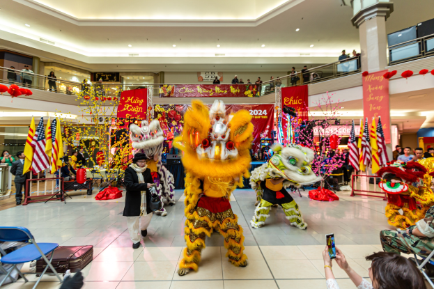Ein lebhaftes chinesisches Neujahrsfest in einem Einkaufszentrum mit Menschen in Kostümen, die tanzen, anderen, die auf Stühlen sitzen und Handys halten, mit Fahnen, Bannern, Dekorationsgegenständen, Geländern, Säulen, Deckenleuchten und einem Wasserzeichen unten.