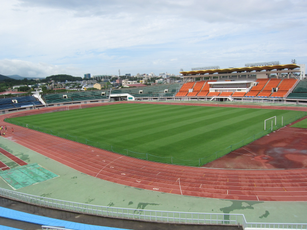 Großes Stadion mit einem Fußballfeld, umgeben von Gebäuden, Bäumen, Hügeln und einem klaren blauen Himmel, mit saftig grünem Gras und ein paar Menschen.