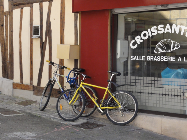 Zwei Fahrräder nebeneinander auf einem Gehweg vor einem Laden mit einer Glaswand und Text, mit einem Objekt an der Wand links von den Fahrrädern.