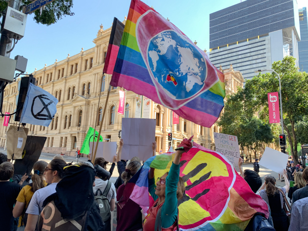 Eine Gruppe von Menschen mit Fahnen und Schildern vor einem Gebäude, umgeben von Bäumen, Laternenmasten, Verkehrszeichen und Texttafeln, die an einer Schwulenparade in Melbourne teilnehmen.
