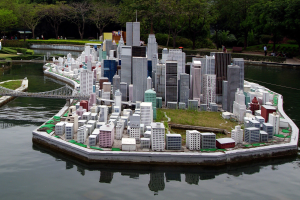 Ein detailliertes Miniaturmodell einer Stadtlandschaft umgeben von Wasser, mit Gebäuden, Grünflächen, einer Brücke, Menschen und Bäumen vor einem Himmel als Hintergrund.