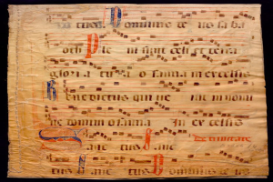 Ein illuminiertes Manuskript mit Musiknotation auf einem Blatt Papier vor dunklem Hintergrund, gefüllt mit Text und musikalischen Symbolen, die ein komplexes Design bilden.