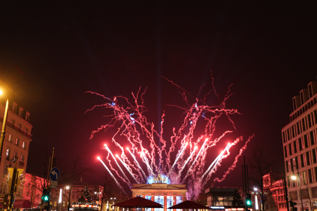 Eine belebte Stadtstraße in Berlin an Silvester, voller Menschen, Fahrzeuge und erleuchtet von Feuerwerk und Gebäudelichtern, die eine festliche Atmosphäre schaffen.