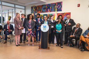 Gruppe von Menschen vor einem Rednerpult mit einem Mikrofon und einer Tafel mit der Aufschrift "Sancho-Stadtrat tritt dem Stadtrat bei", mit sitzenden und stehenden Zuschauern, Fahnen, einem Feuerlöscher und Deckenleuchten im Hintergrund.