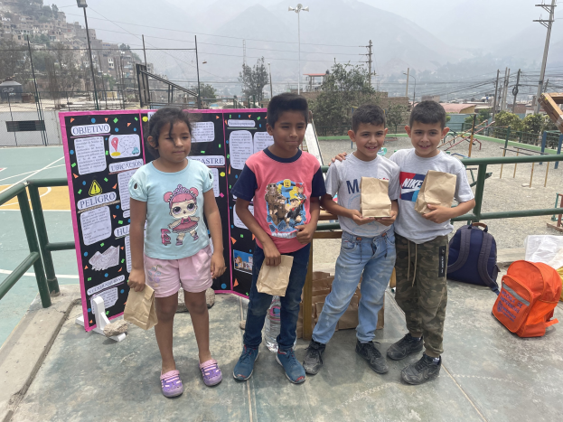 Gruppe von Kindern auf einem Basketballfeld mit Papierbeuteln in den Händen, mit Beuteln und Gegenständen auf der rechten Seite und einer Tafel mit Text auf der linken Seite, vor einem Hintergrund aus Zäunen, Pfählen, Straßenlaternen, Bäumen, Gebäuden, Hügeln und Himmel.