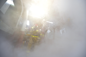 Eine Gruppe von Feuerwehrmännern in Helmen und Schutzausrüstung steht in einem raucherfüllten Raum, umgeben von verstreuten Gegenständen.