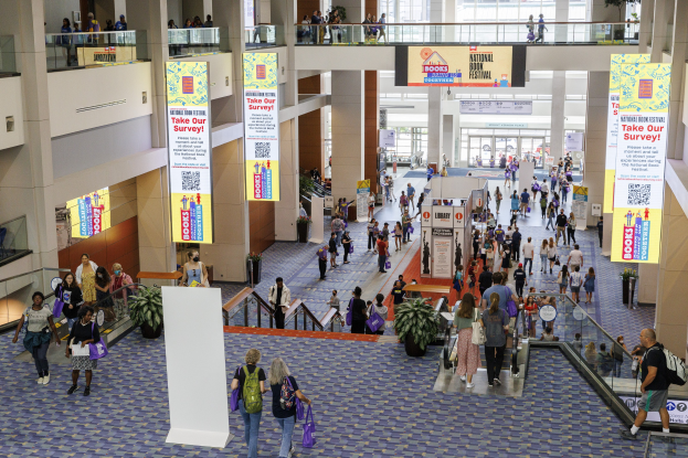 Eine große Gruppe von Menschen geht durch ein großes Gebäude mit Bannern, Topfpflanzen, Rolltreppen, Geländern, Säulen, Texttafeln und Glastüren, einige tragen Taschen, wahrscheinlich besuchen sie das National Book Festival.