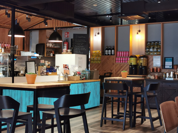 Inneneinrichtung eines Restaurants mit Holztischen, Stühlen, einer Topfpflanze auf einem Tisch, einer Schiefertafelmenü, einem Kühlschrank, einem Feuerlöscher, Regalen mit Flaschen, Schränken mit Gegenständen, einem Rahmenfoto an der Wand und Deckenleuchten.