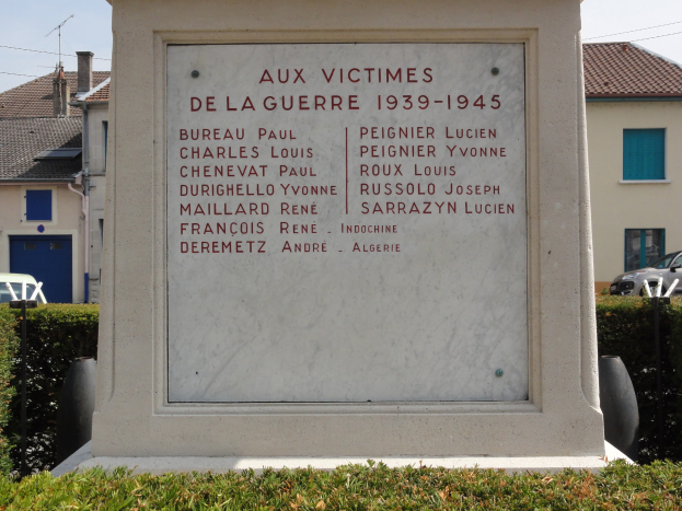 Ein Denkmal für die Opfer des Krieges von 1939–1945 steht vor einem Gebäude mit der Aufschrift "Aux Victimes de la Guerre 1939-1945", umgeben von Pflanzen, Fahrzeugen, Strommasten und nahegelegenen Häusern unter einem bewölkten Himmel.