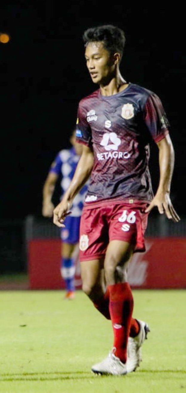 Ein Mann in einem rotbraunen Hemd und roten Shorts, der auf einem Feld Fußball spielt, mit einer anderen Person im Hintergrund und einer Tafel mit Schrift.