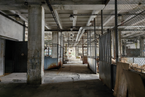 Ein leerer, verlassener Fabrikraum mit großen Fenstern, Säulen, einer Tür, einer Decke mit Deckenleuchten, Metallpfosten, einem Maschendrahtzaun, verstreuten Gegenständen auf dem Boden und einer Wand, die einen Eindruck von Vernachlässigung vermittelt.