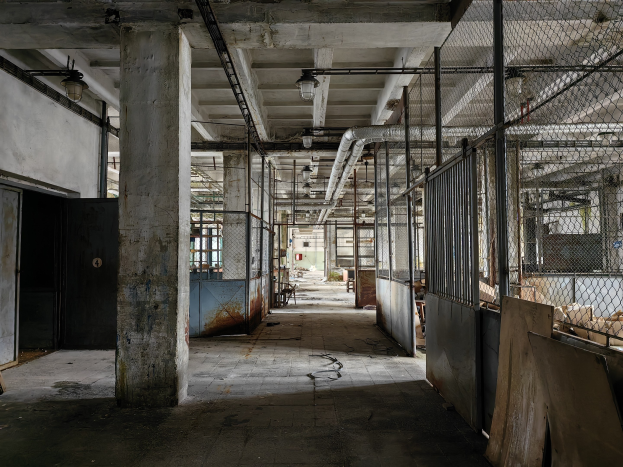 Ein leerer, verlassener Fabrikraum mit großen Fenstern, Säulen, einer Tür, einer Decke mit Deckenleuchten, Metallpfosten, einem Maschendrahtzaun, verstreuten Gegenständen auf dem Boden und einer Wand, die einen Eindruck von Vernachlässigung vermittelt.