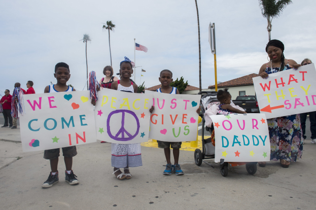 Gruppe von Kindern mit Schildern 'Wir kommen in Frieden, gebt uns unseren Daddy' auf einer Straße mit einer Frau, die einen Kinderwagen schiebt, umgeben von Bäumen, Pfählen, einer Fahne, einem Haus, Pflanzen und einem bewölkten Himmel.