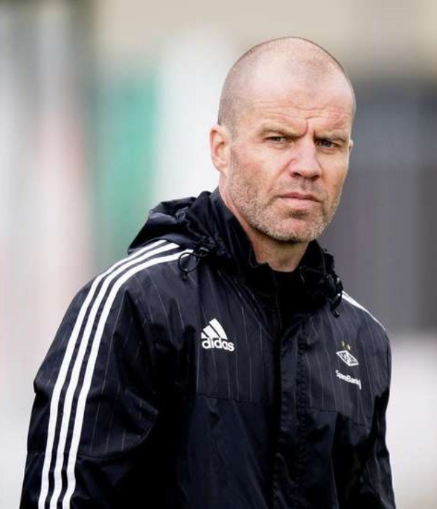 Kahlköpfiger Fussballtrainer in marineblauer Jacke mit weissen Streifen und weissem Kragen auf einem Feld stehend