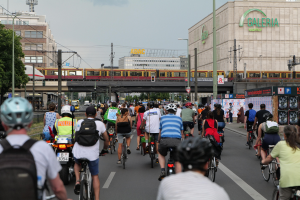 Eine Gruppe von Menschen, die Fahrräder auf einer Straße mit hohen Gebäuden fährt, wobei einige Helme und Taschen tragen und ein Zug auf einem Bahngeleise im Hintergrund zu sehen ist.