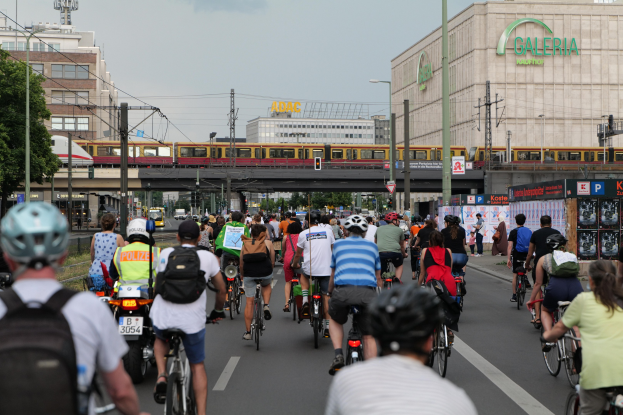 Eine Gruppe von Menschen, die Fahrräder auf einer Straße mit hohen Gebäuden fährt, wobei einige Helme und Taschen tragen und ein Zug auf einem Bahngeleise im Hintergrund zu sehen ist.