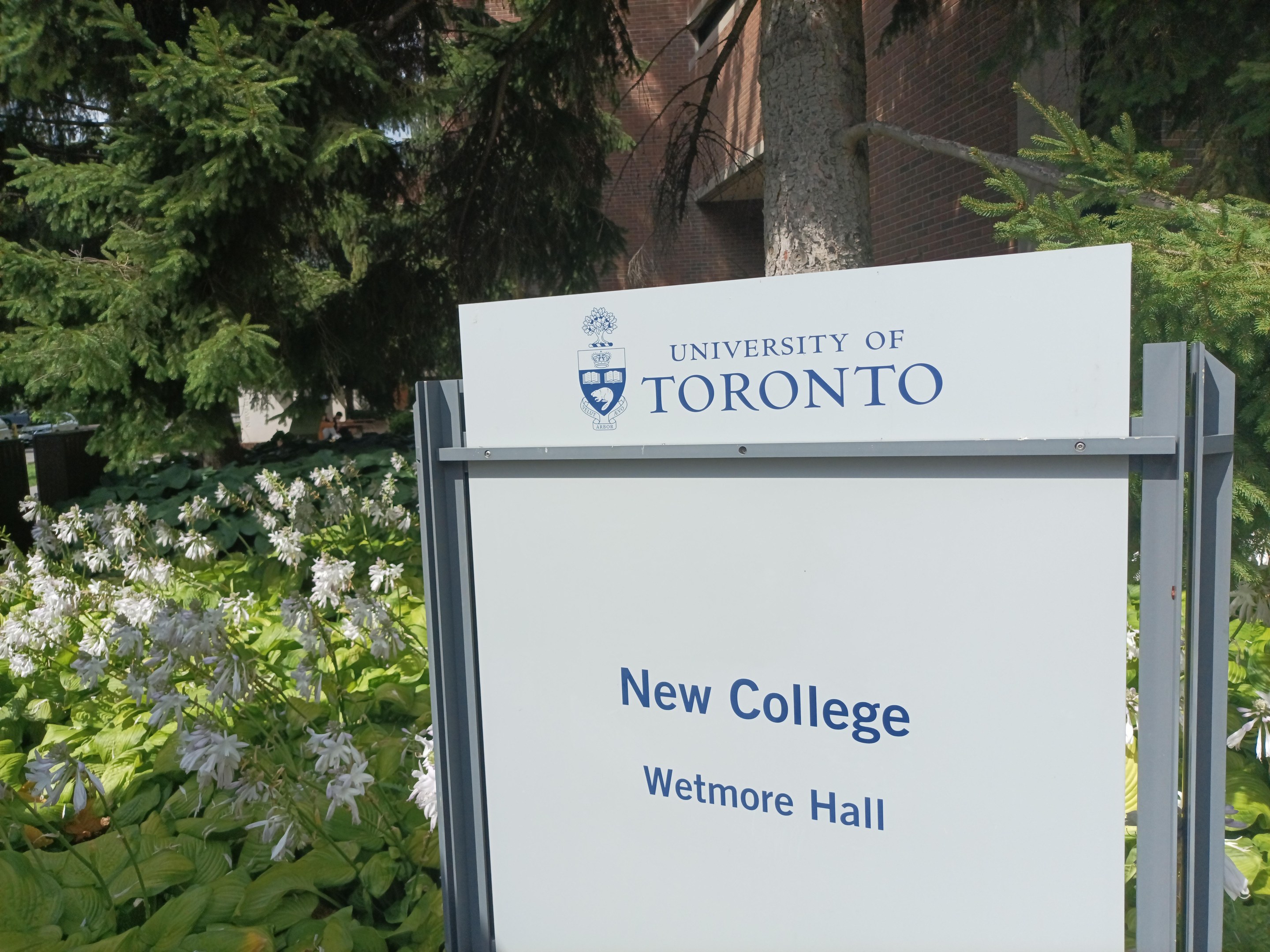 Außenansicht des neuen Colleges der Universität Toronto an Wetmore Hall mit einem Schild vor dem Gebäude, umgeben von grünem Gras, bunten Blumen, hohen Bäumen und einem hellblauen Himmel.