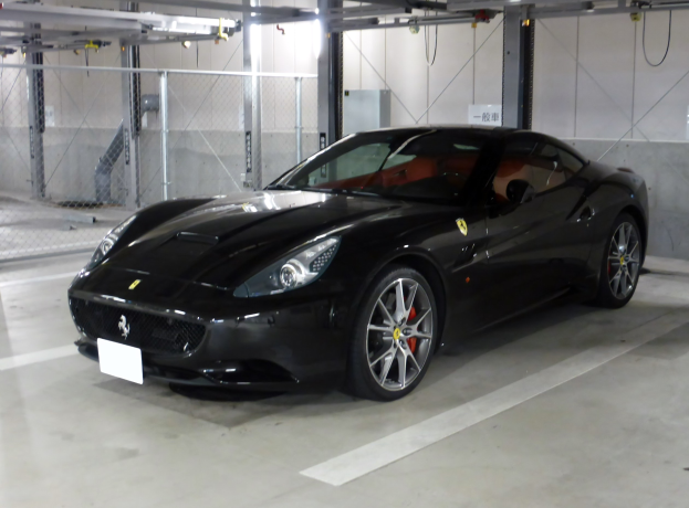 Schwarzer Ferrari California in einer Parkgarage mit einer Wand im Hintergrund, Maschendrahtzaun links und Deckenbeleuchtung.