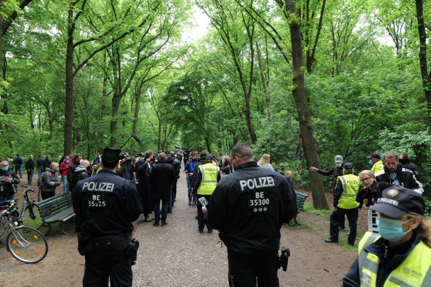 Eine Gruppe von Polizeibeamten steht vor einer Menschenmenge, einige tragen Mützen und Masken, mit Fahrrädern und einer Bank im Vordergrund und Bäumen und Himmel im Hintergrund, während einer Anti-Terror-Demonstration in Berlin.
