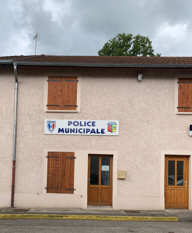 Polizeigebäude mit einem Schild, Fenstern, Türen, einer Namensplakette, einem Rohr, einem Baum und einem bewölktem Himmel im Hintergrund.