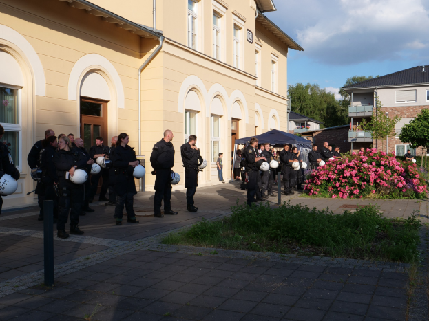 Gruppe von Polizeibeamten vor einem Gebäude mit Fenstern und Türen stehend, Helme haltend, in der Nähe eines Zeltes mit Pflanzen, eines Straßenmastes, Bäumen und einem bewölkten Himmel.