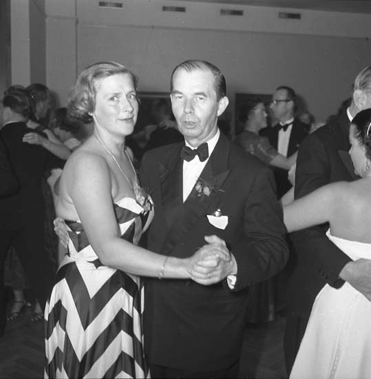 Ein Mann in einem schwarzen Blazer, weißem Hemd und schwarzen Hosen tanzt mit einer Frau in unbekannter Kleidung auf einer formellen Veranstaltung, indem sie sich an den Händen halten und zur Musik schwenken, nahe an einer Wand.