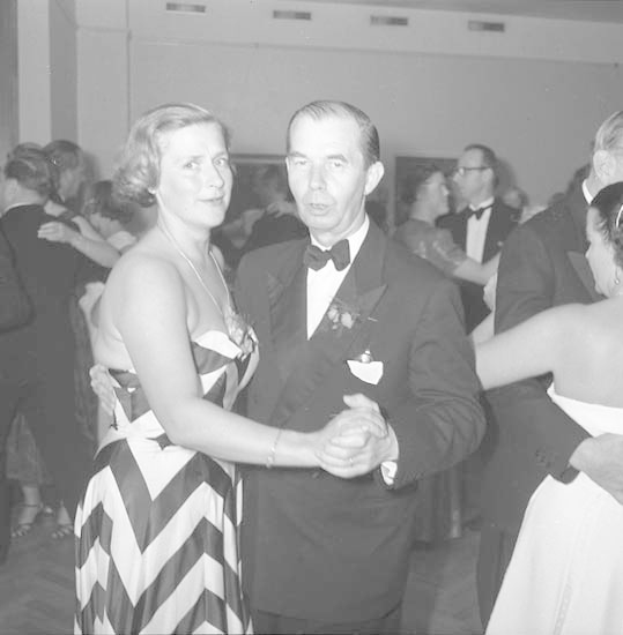 Ein Mann in einem schwarzen Blazer, weißem Hemd und schwarzen Hosen tanzt mit einer Frau in unbekannter Kleidung auf einer formellen Veranstaltung, indem sie sich an den Händen halten und zur Musik schwenken, nahe an einer Wand.